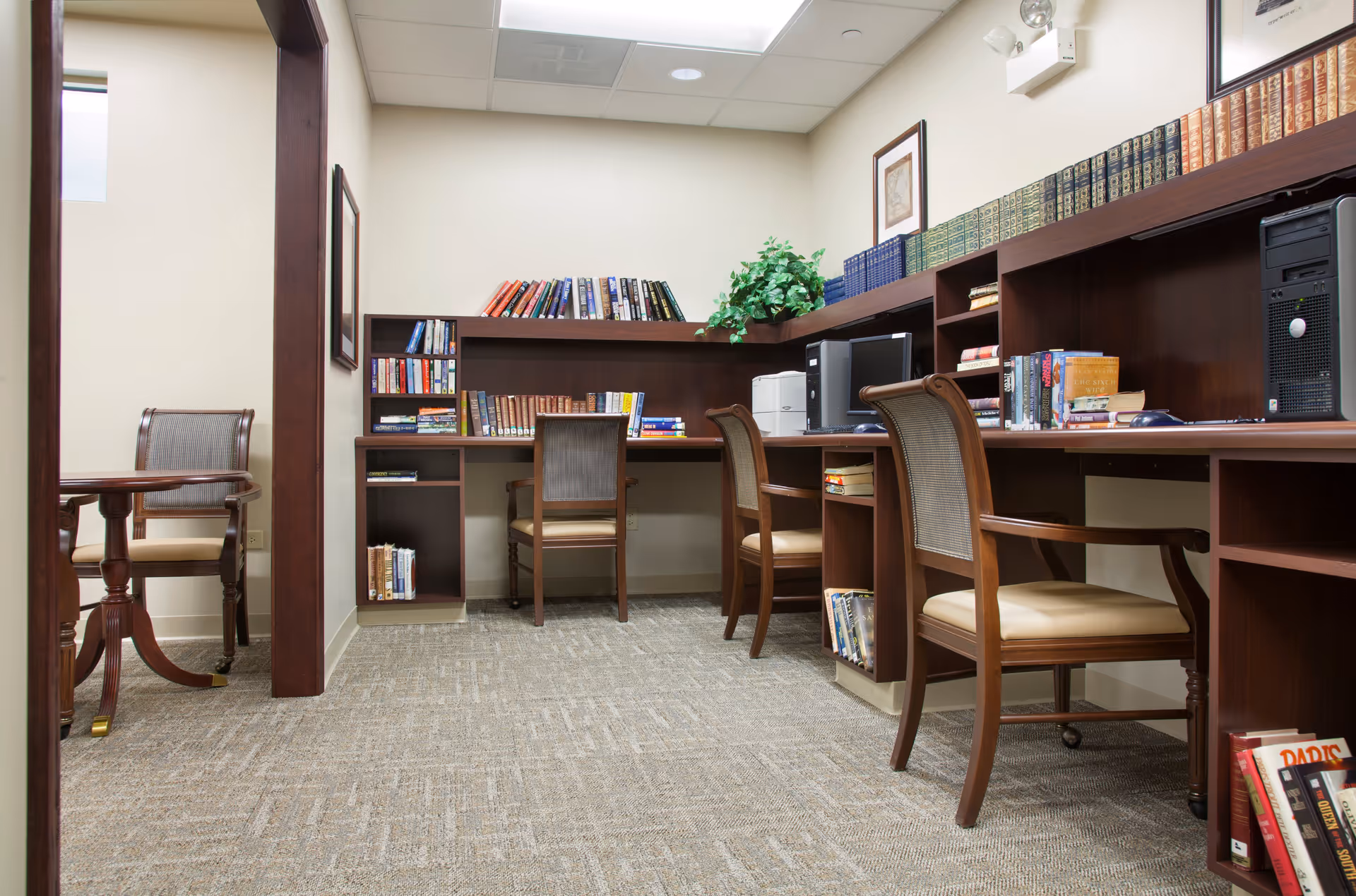 A quiet room with wooden desks and chairs arranged along the walls, featuring bookshelves filled with books, a computer, a printer, and a potted plant. The room has beige walls and carpeted flooring, with framed artwork on the walls.