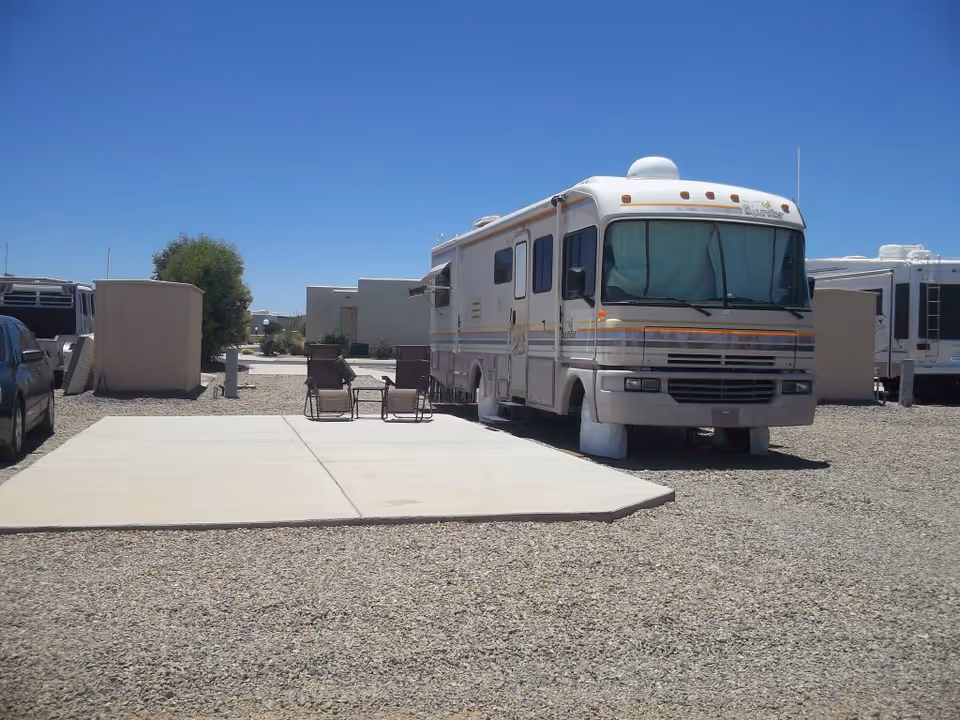An outdoor RV parking area at Vista View Resort with a large white RV parked on a gravel lot next to a concrete pad with two brown chairs and a small table. Other vehicles and RVs are visible in the background under a clear blue sky.