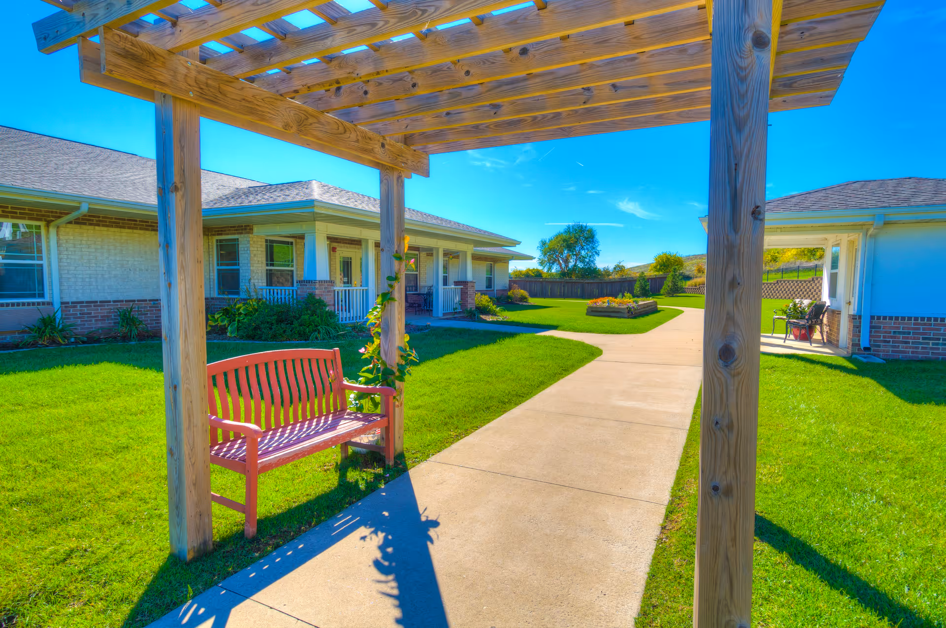 Outdoor courtyard area at River Valley Place of Fort Madison featuring a wooden pergola with a red bench underneath, a concrete walkway, green grass, flower beds, and single-story buildings with porches in the background under a clear blue sky.