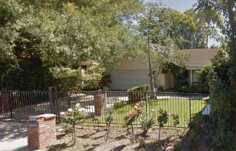 Front exterior view of a single-story house with a two-car garage, surrounded by trees and a garden with rose bushes behind a metal fence with brick pillars.