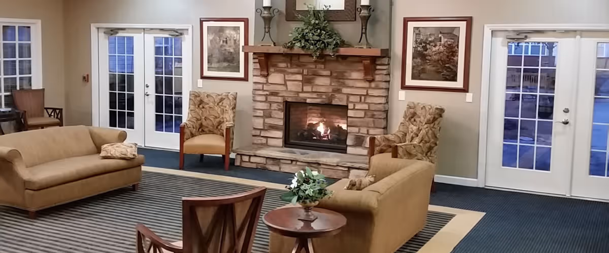 A cozy senior living facility common area with a stone fireplace in the center, two beige sofas facing each other, two patterned armchairs, a small round wooden table with a plant, framed artwork on the walls, and double glass doors on either side of the fireplace.