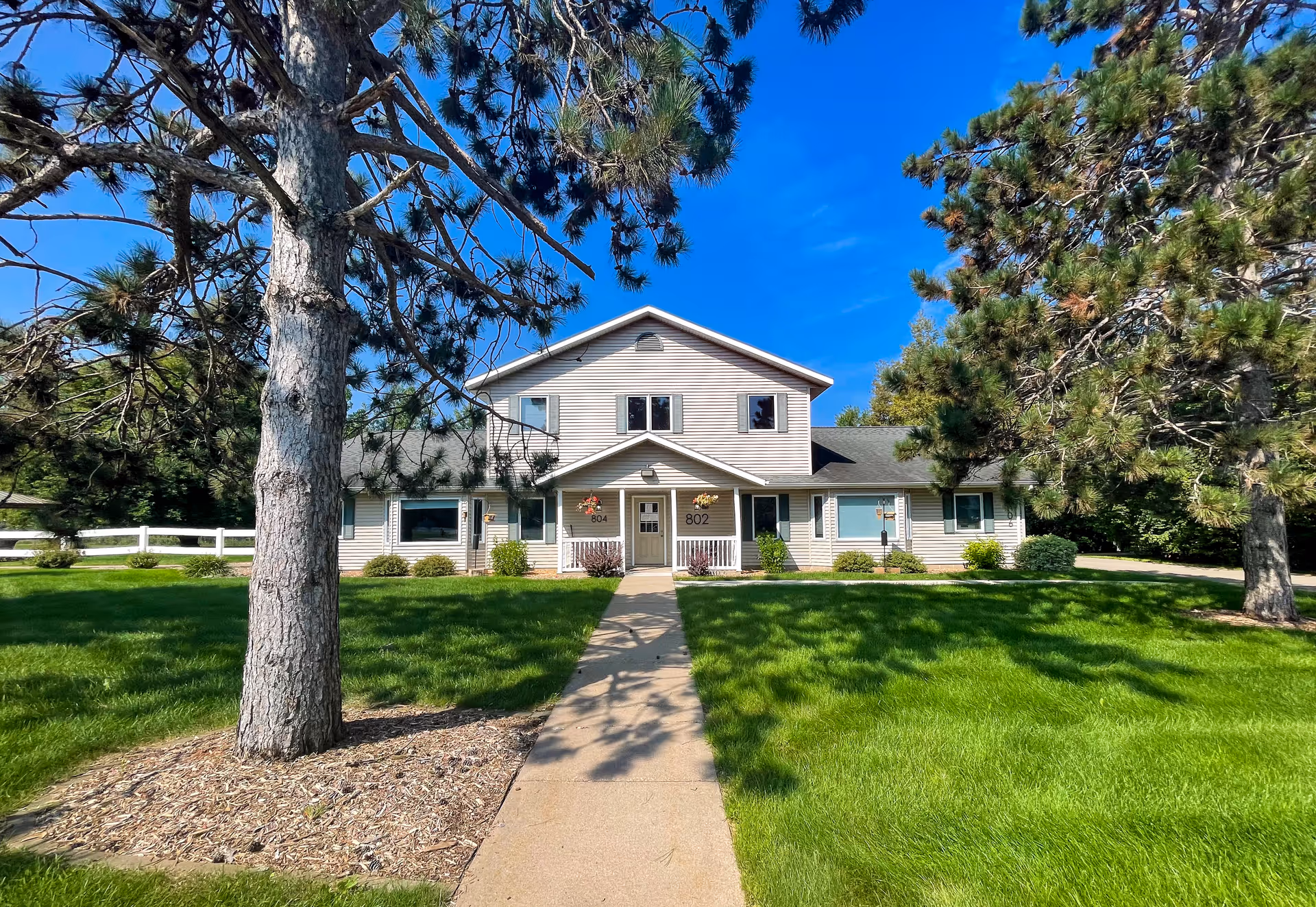 Front exterior of a two-story residential building with a central sidewalk, green lawn, and large pine trees.