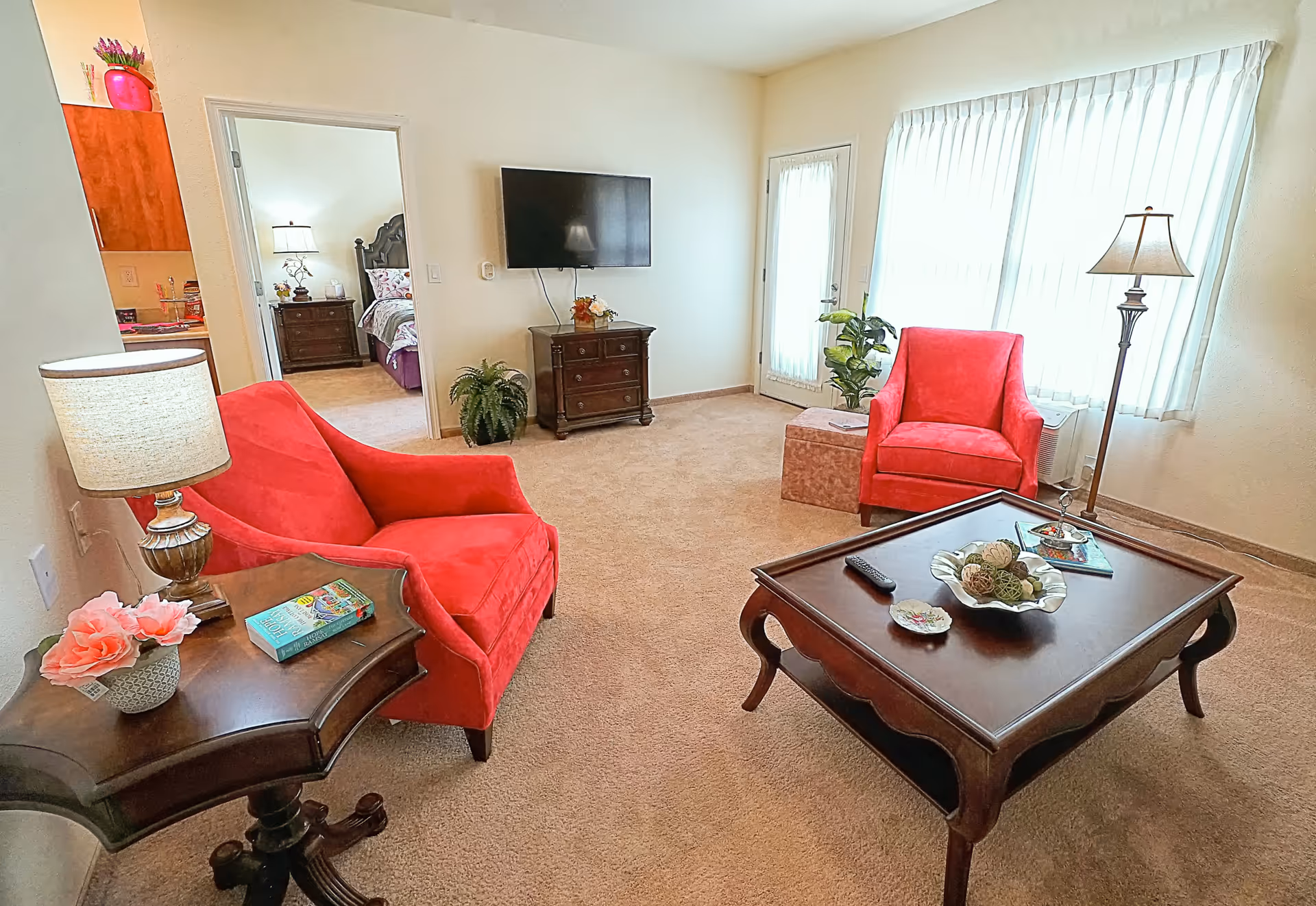 Bright living room with two red armchairs, a wooden coffee table, a wall-mounted TV, and a doorway leading to a bedroom.