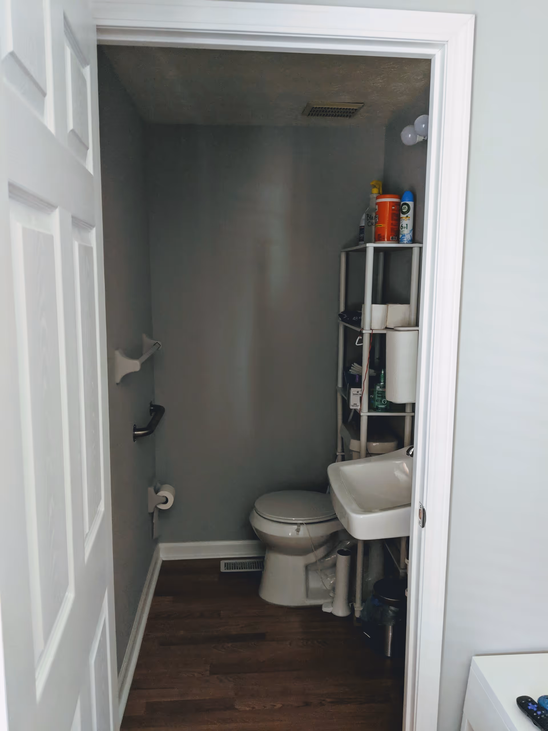 Small bathroom with a white toilet and a white sink. There is a shelving unit next to the sink holding various cleaning supplies and paper towels. The walls are painted gray and the floor is wooden. A white door is partially open, revealing the bathroom interior.