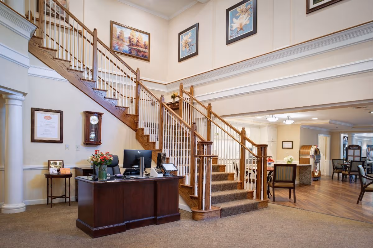 Lobby of a senior living facility with a wooden reception desk, a grand staircase, and a seating/dining area in the background.