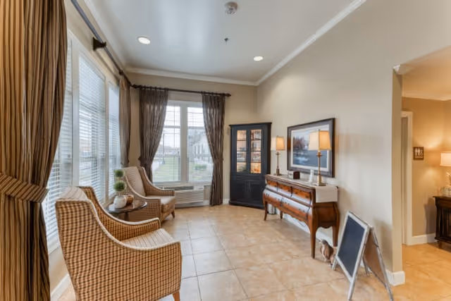 A bright and cozy sitting area in a senior living facility with two cushioned armchairs near large windows with brown curtains. A small round table with decorative items is placed between the chairs. Across from the chairs is a wooden sideboard with two table lamps and a framed picture above it. A black display cabinet stands in the corner, and a small chalkboard sign is propped against the wall near the entrance to another room.