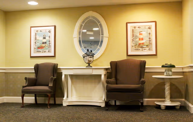 Two upholstered armchairs flank a white console table beneath an oval mirror and framed lighthouse prints in a softly lit seating area.
