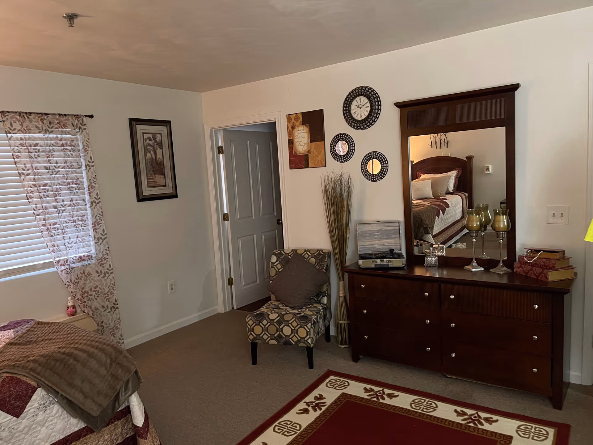 A cozy bedroom with a bed partially visible on the left covered with a quilt and a brown blanket. There is a window with floral curtains and blinds, a patterned armchair with a cushion, and a wooden dresser with a large mirror. On the dresser are decorative items including three candle holders, a small box, and a stack of books. The wall above the dresser features a clock and two round mirrors. The door to another room is open.