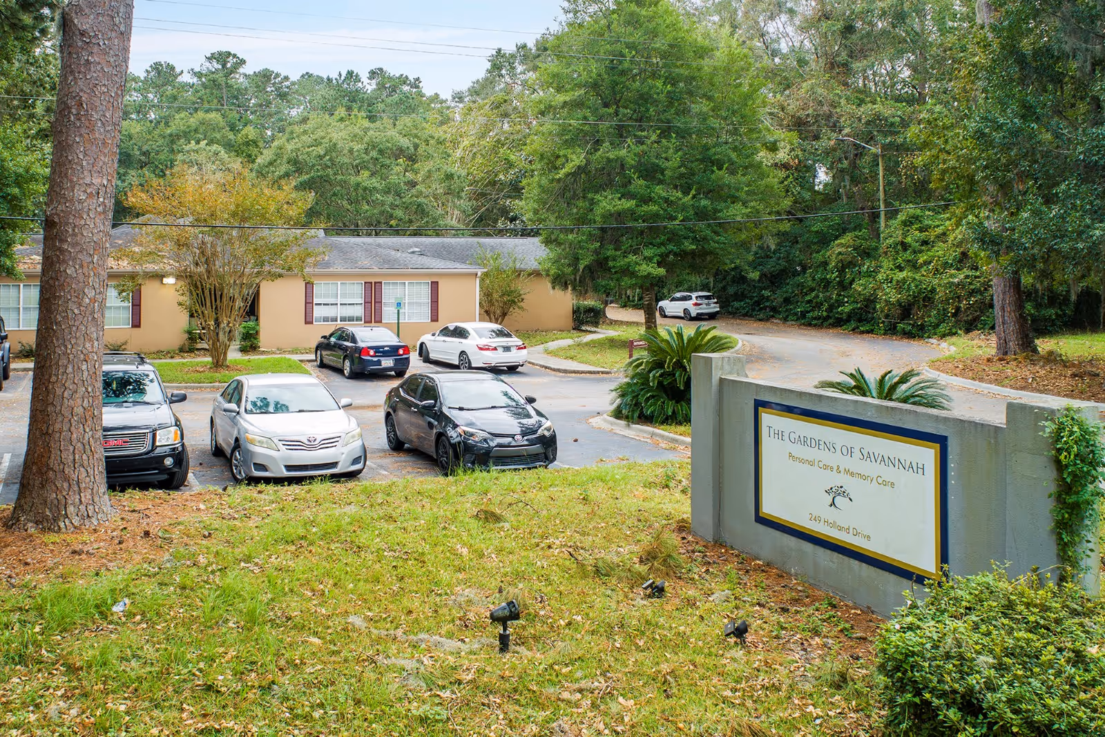 Parking lot and entrance sign for The Gardens of Savannah with cars in front of a single-story building and surrounding trees.