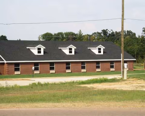 Brick senior living center exterior with dormer windows along a grassy roadside.