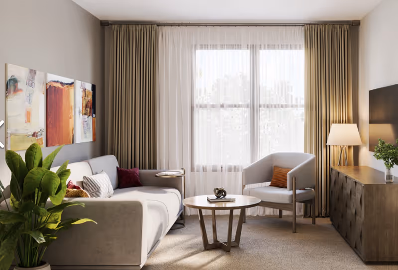 Sunlit living room with a sofa, armchair, round coffee table, console with lamp, artwork, and a large curtained window.
