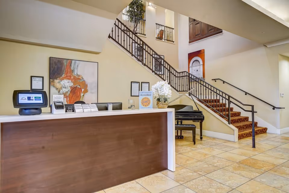 Lobby reception desk with a computer terminal, floral arrangement, grand piano, and a carpeted staircase.