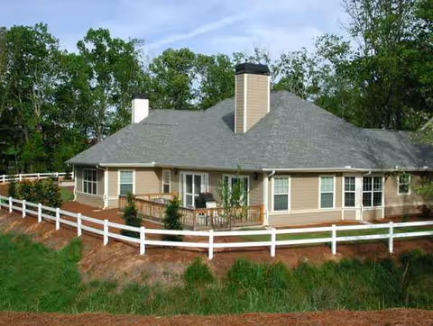 Single-story beige senior living building with a gray roof, white fence, and surrounding trees.