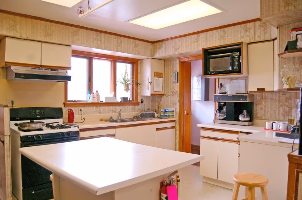 A bright kitchen with a white island countertop in the center, a gas stove with a pan on it, a double sink under a window with some plants and cleaning supplies on the windowsill, a microwave above a coffee maker, and cabinets with a beige and brown color scheme. There is a wooden stool near the counter and patterned wallpaper on the walls.