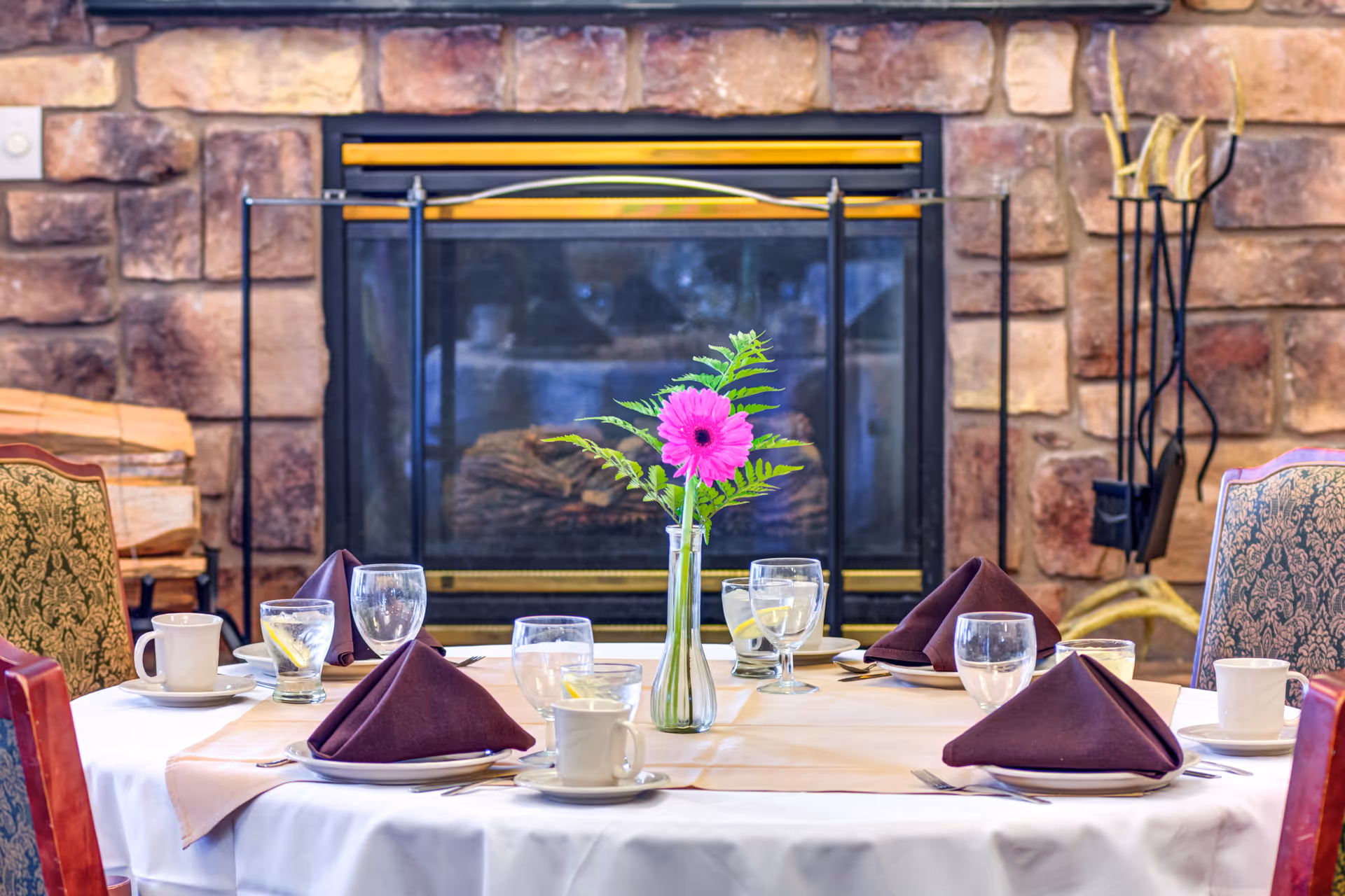 Round dining table set for four with folded napkins, glassware and a pink flower centerpiece in front of a stone fireplace.