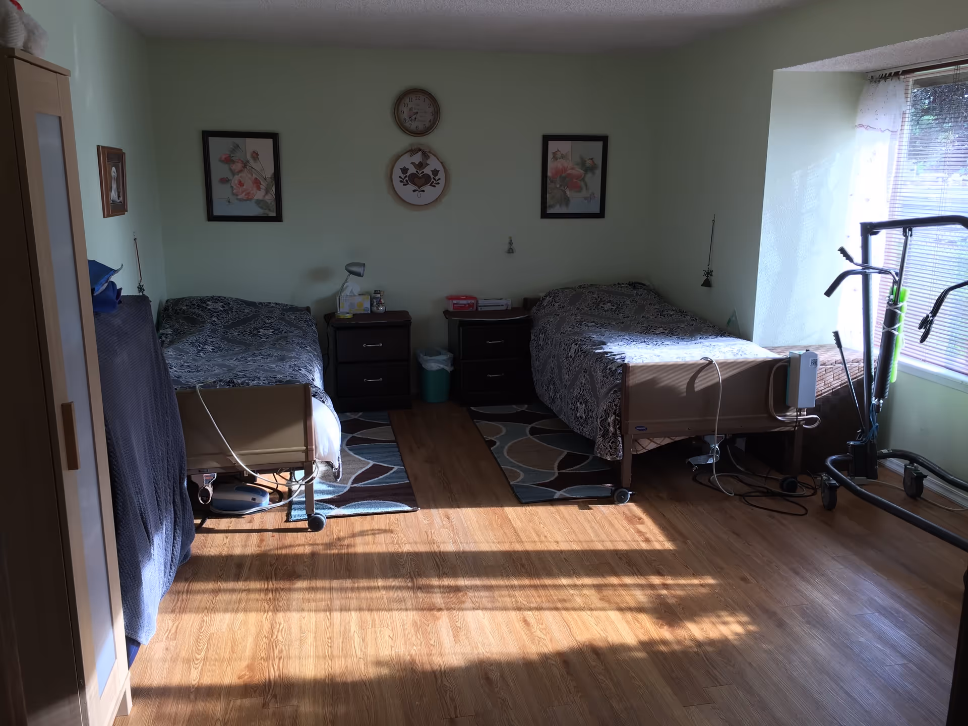 A sunny shared bedroom with two hospital-style single beds, nightstands, framed artwork, a window, and exercise equipment.