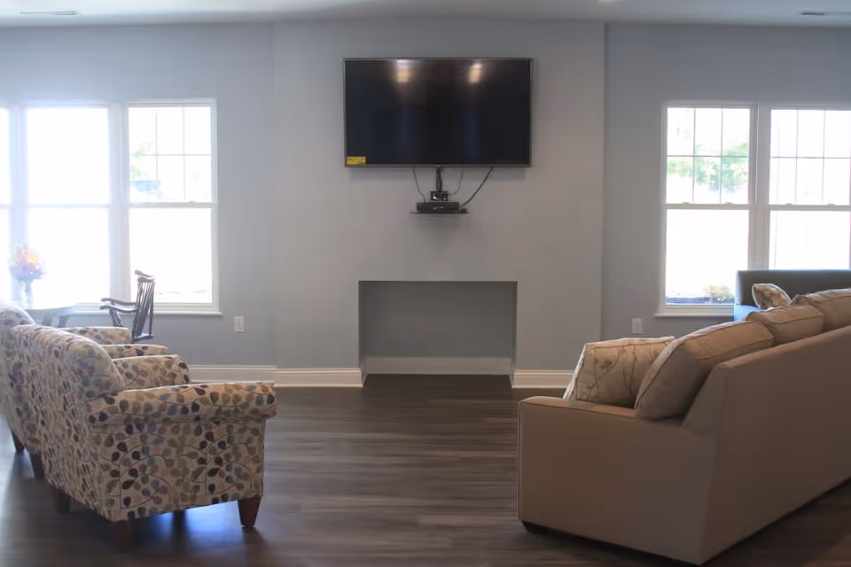 A bright living room with two large windows on either side of a wall-mounted flat screen TV. The room features a patterned armchair, a beige sofa with decorative pillows, and a wooden chair near the window. The floor is dark wood, and the walls are painted light gray.