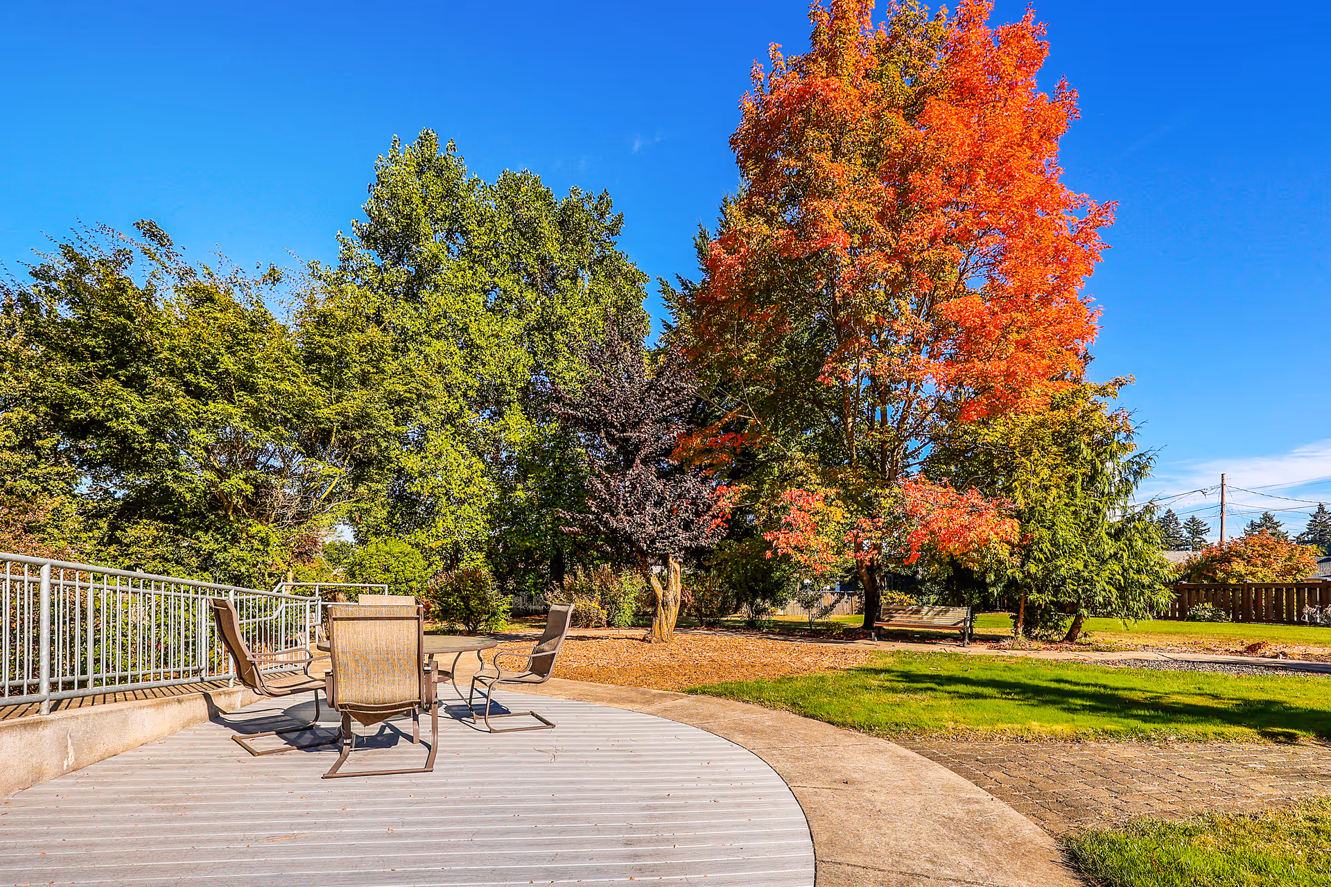 Outdoor patio area with four chairs around a circular table on a wooden deck, surrounded by green grass and trees, including one with bright red autumn leaves under a clear blue sky.