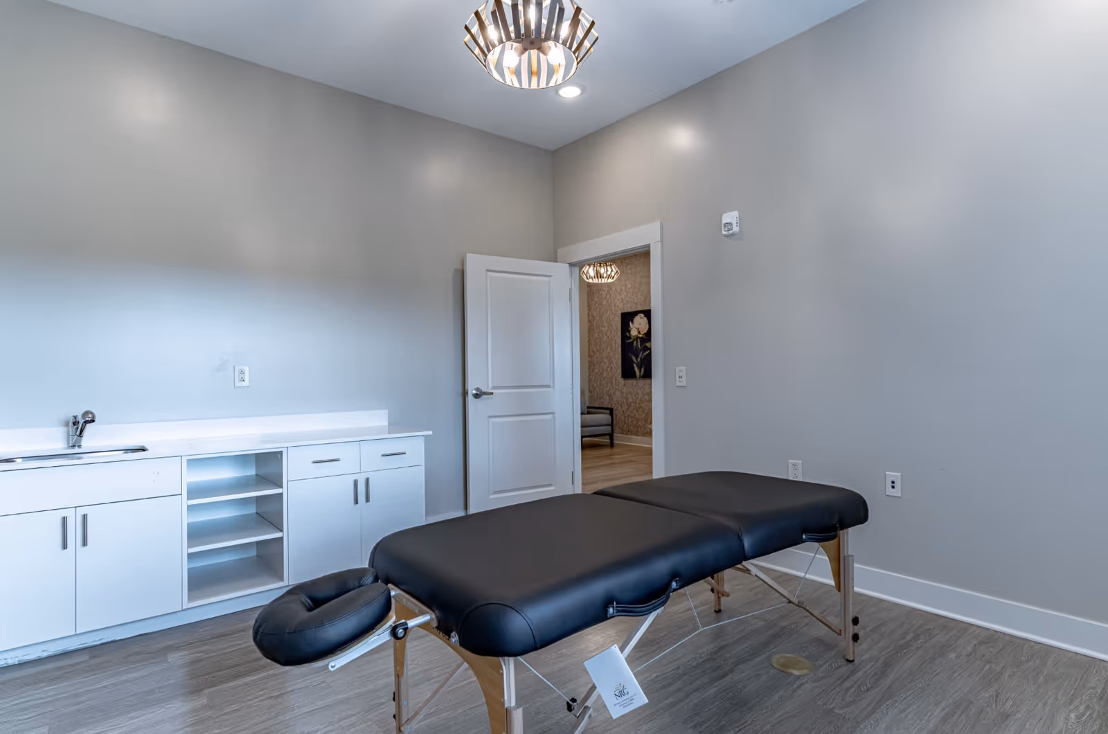 A treatment room with a black massage table, white cabinetry with a sink, and an open door to an adjacent room.