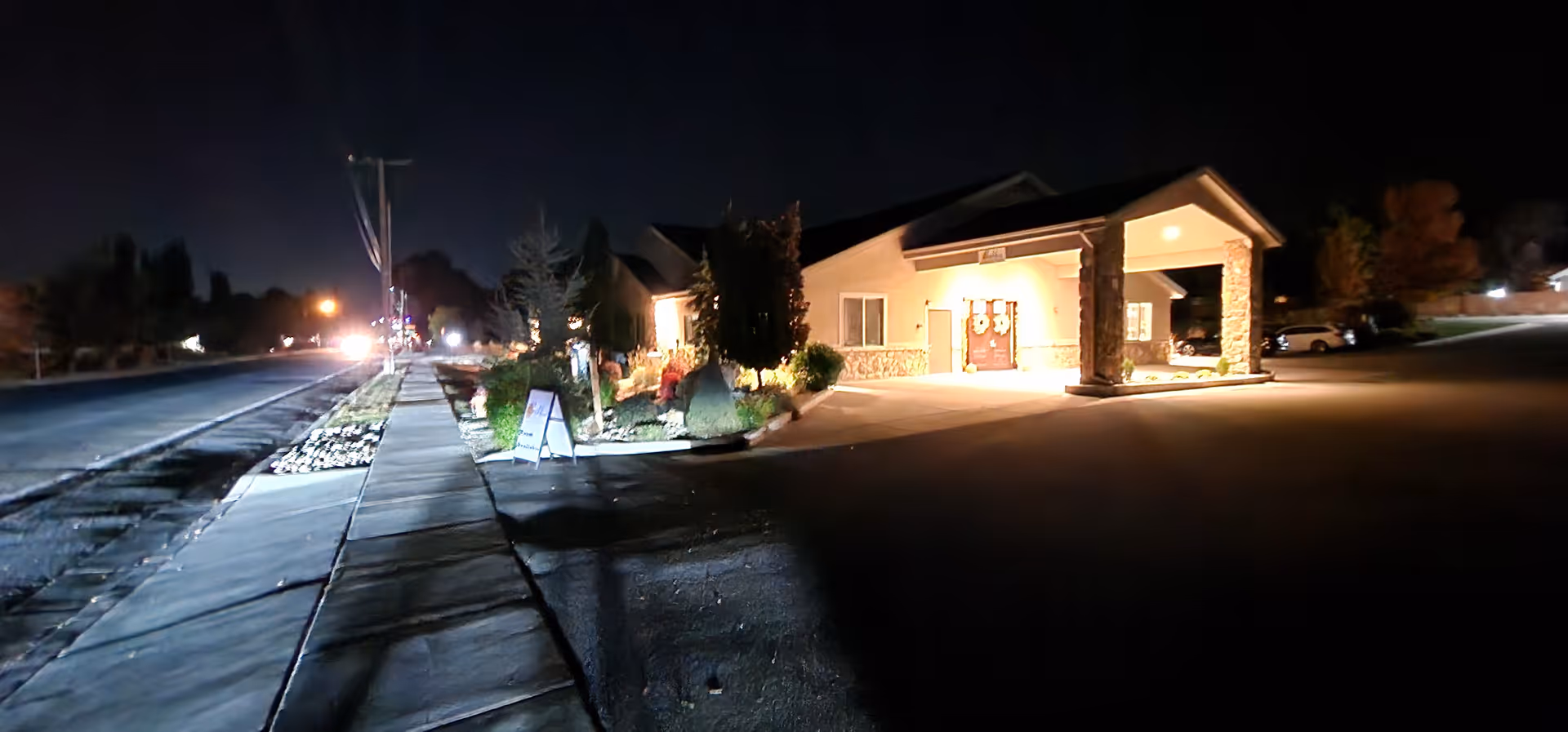 Nighttime view of the front exterior of a single-story building with a covered entrance supported by stone pillars, illuminated by outdoor lights. A sidewalk runs along the left side of the building, and there are some bushes and landscaping near the entrance. The surrounding area is dark with a few distant lights visible.