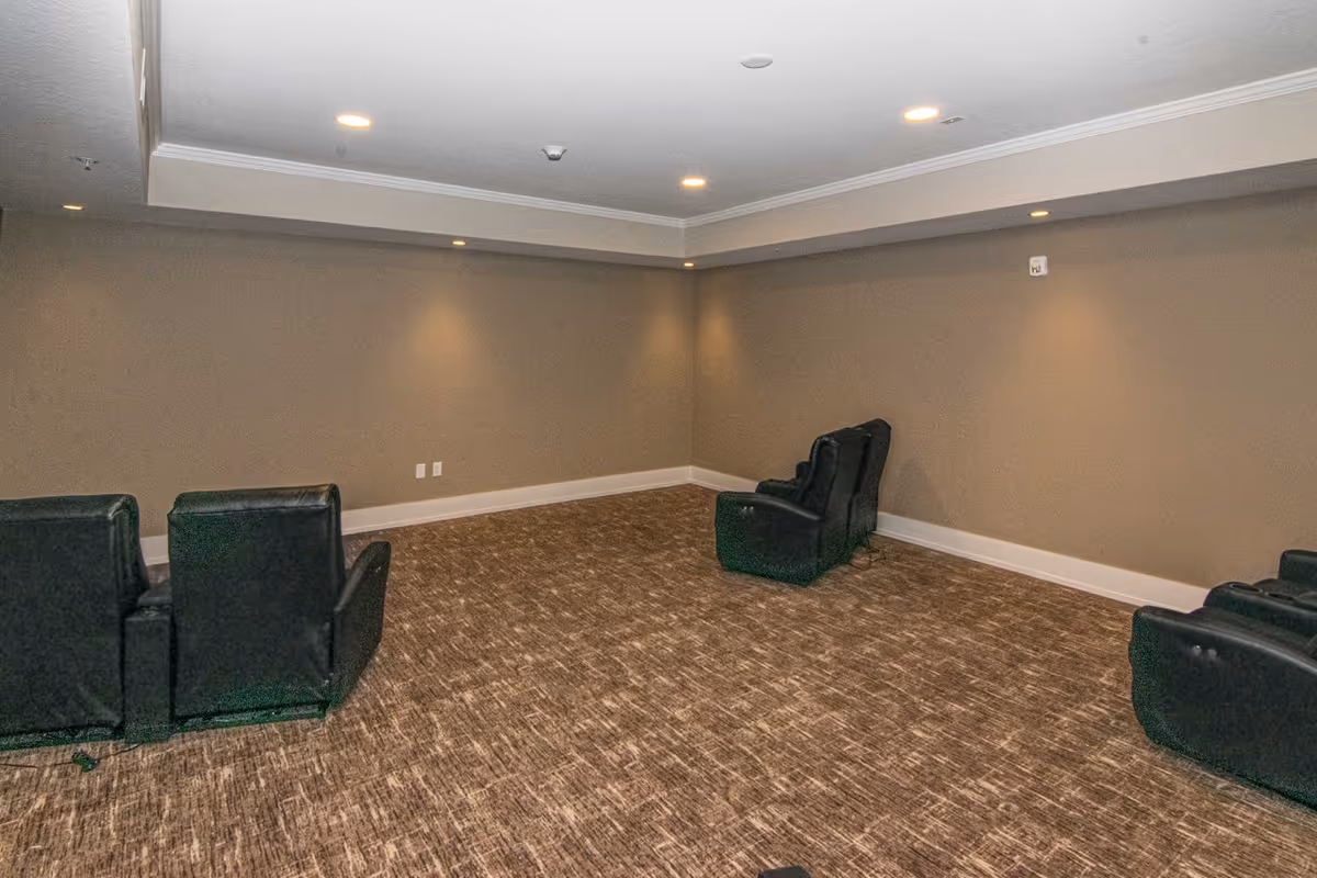 A small room with beige walls and carpeted floor featuring four black recliner chairs arranged in two pairs facing each other. The ceiling has recessed lighting and a white crown molding along the edges.