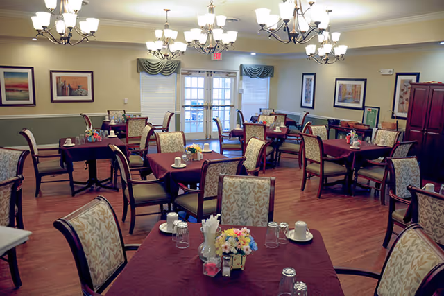 Communal dining room with multiple tables set with place settings, chandeliers overhead, and double doors at the far wall.