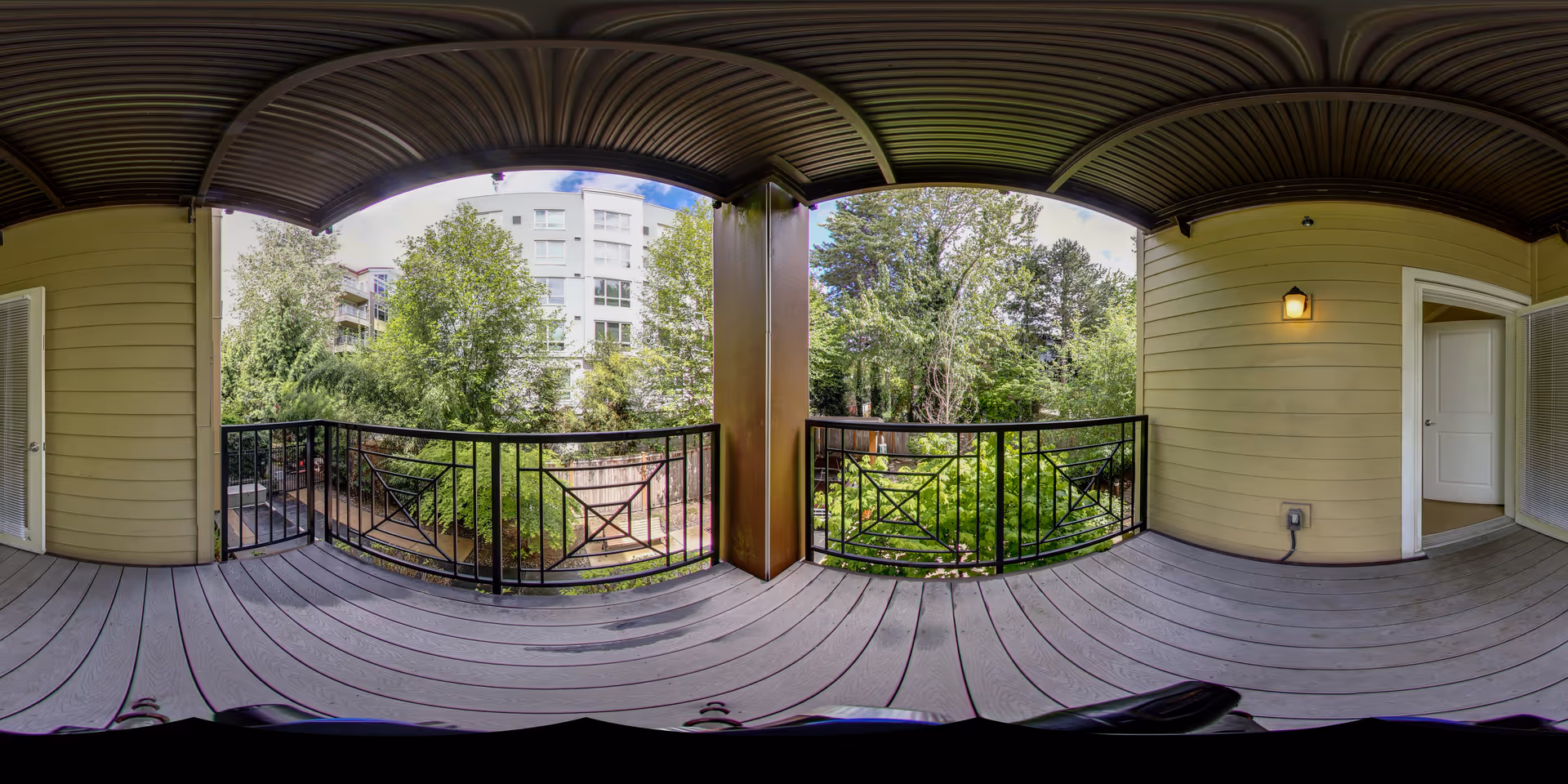 Covered balcony with wooden flooring and black metal railing overlooking a green garden area with trees and shrubs. The balcony has a ceiling light and is attached to a building with yellow siding. An open door leads inside the building.