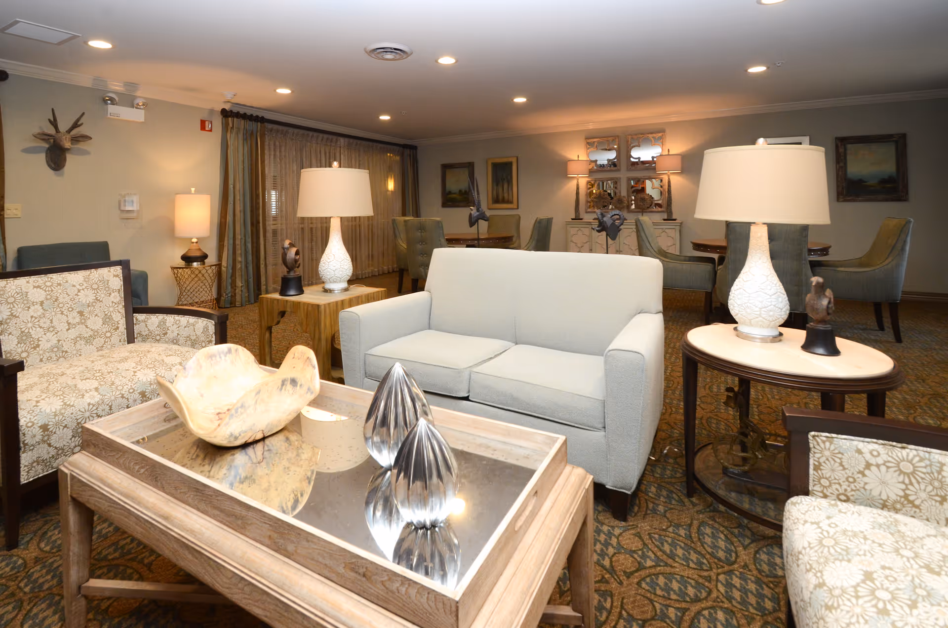 A cozy living room area with a light gray loveseat, two patterned armchairs, and a wooden coffee table with decorative items. There are two side tables with white lamps, and in the background, there are additional chairs, a cabinet with mirrors, framed artwork on the walls, and curtains covering a window.