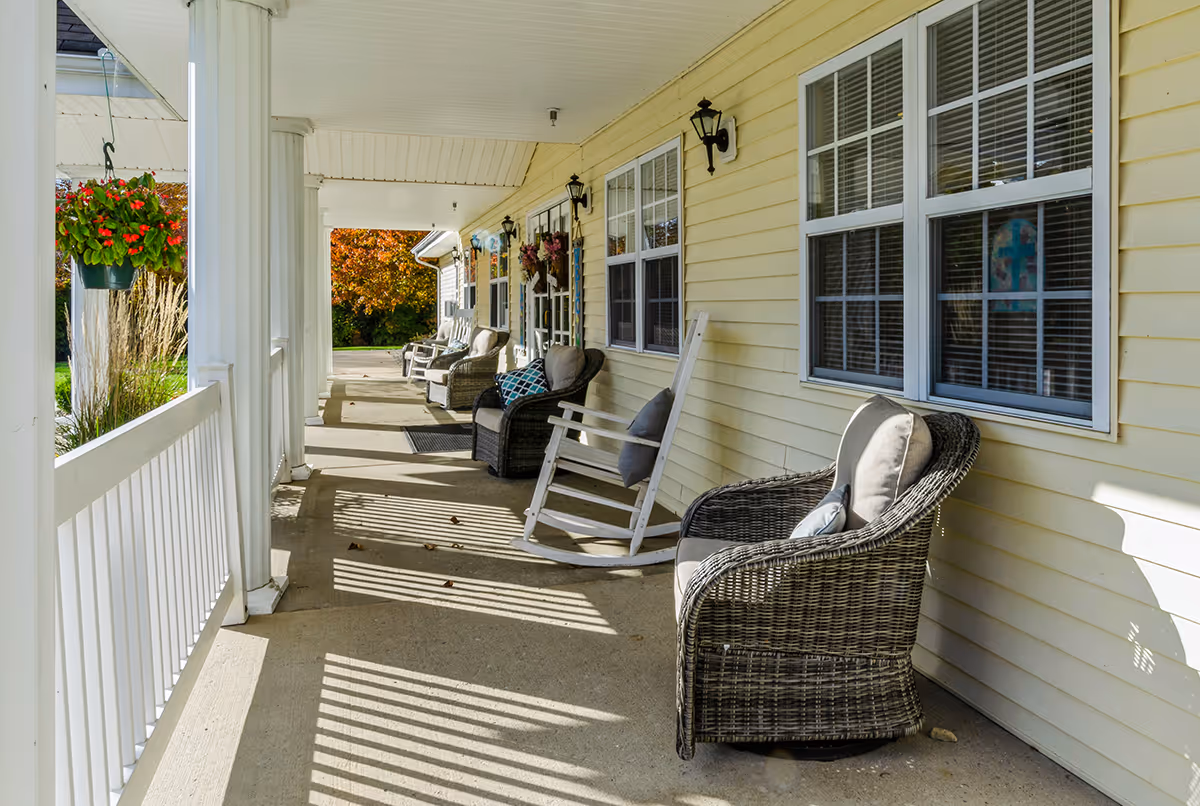 A sunny porch area with several cushioned wicker chairs and a white rocking chair lined up against a yellow siding wall with windows. Hanging flower baskets and outdoor wall lanterns are also visible, with a view of green grass and autumn-colored trees in the background.