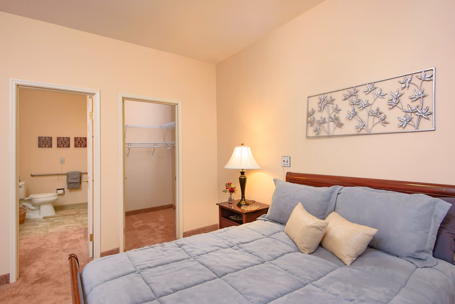 A bedroom with a neatly made bed featuring a light blue comforter and two beige pillows. A wooden nightstand with a lamp and a small vase with flowers is beside the bed. On the wall above the bed is a decorative metal wall art with a leaf design. Two open doorways are visible, one leading to a bathroom with a toilet and towel rack, and the other to a walk-in closet with wire shelving. The walls are painted a soft beige color and the carpet is light brown.