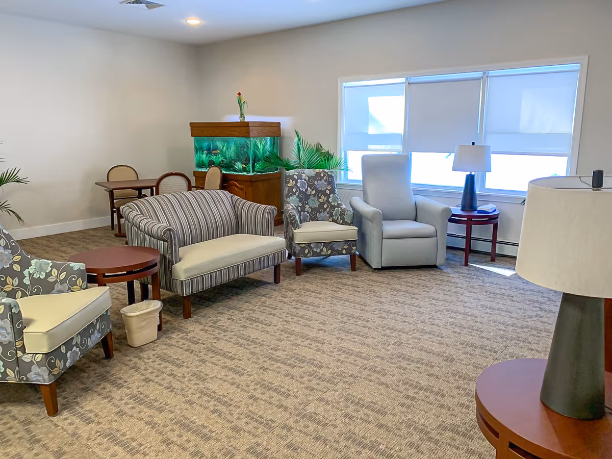 A bright and spacious common area with various seating options including a striped loveseat, floral armchairs, and a gray recliner. There is a large fish tank against the wall, a small round table with a lamp, and a window with blinds letting in natural light. The room has beige carpet and neutral-colored walls.