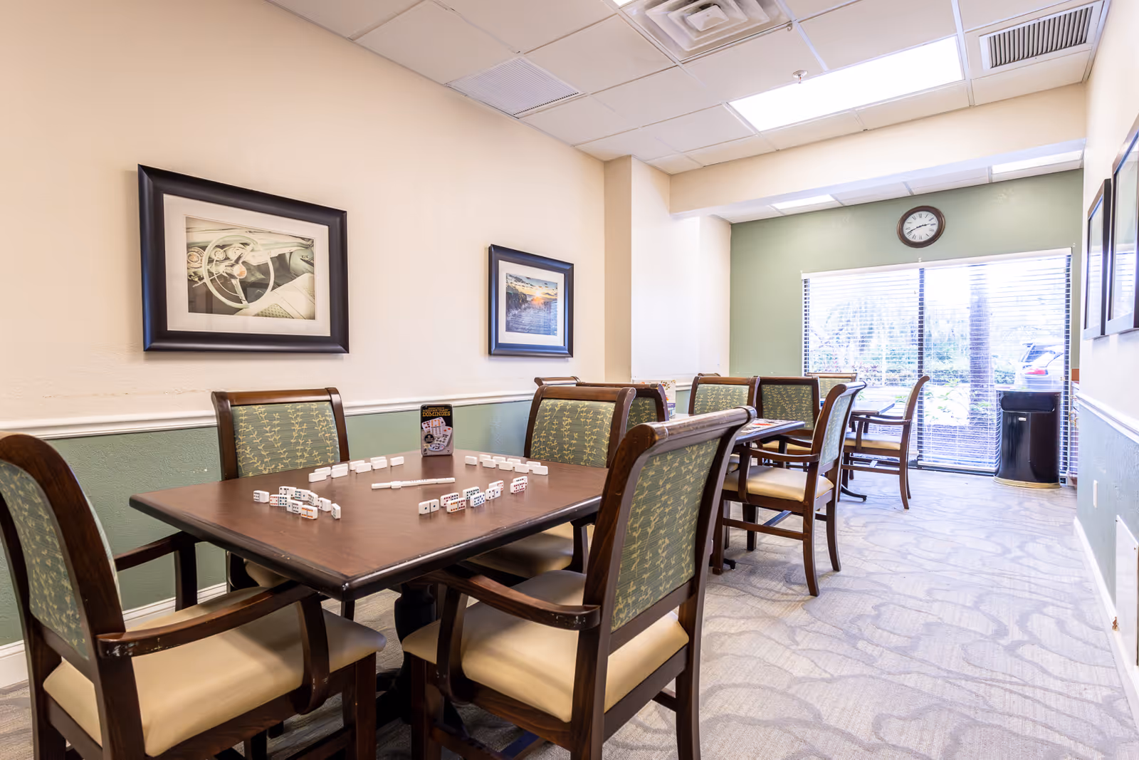Bright communal dining/activity room with several wooden tables and upholstered chairs, a game of dominoes set on the nearest table, framed artwork on the walls, and a clock above a sliding glass door.