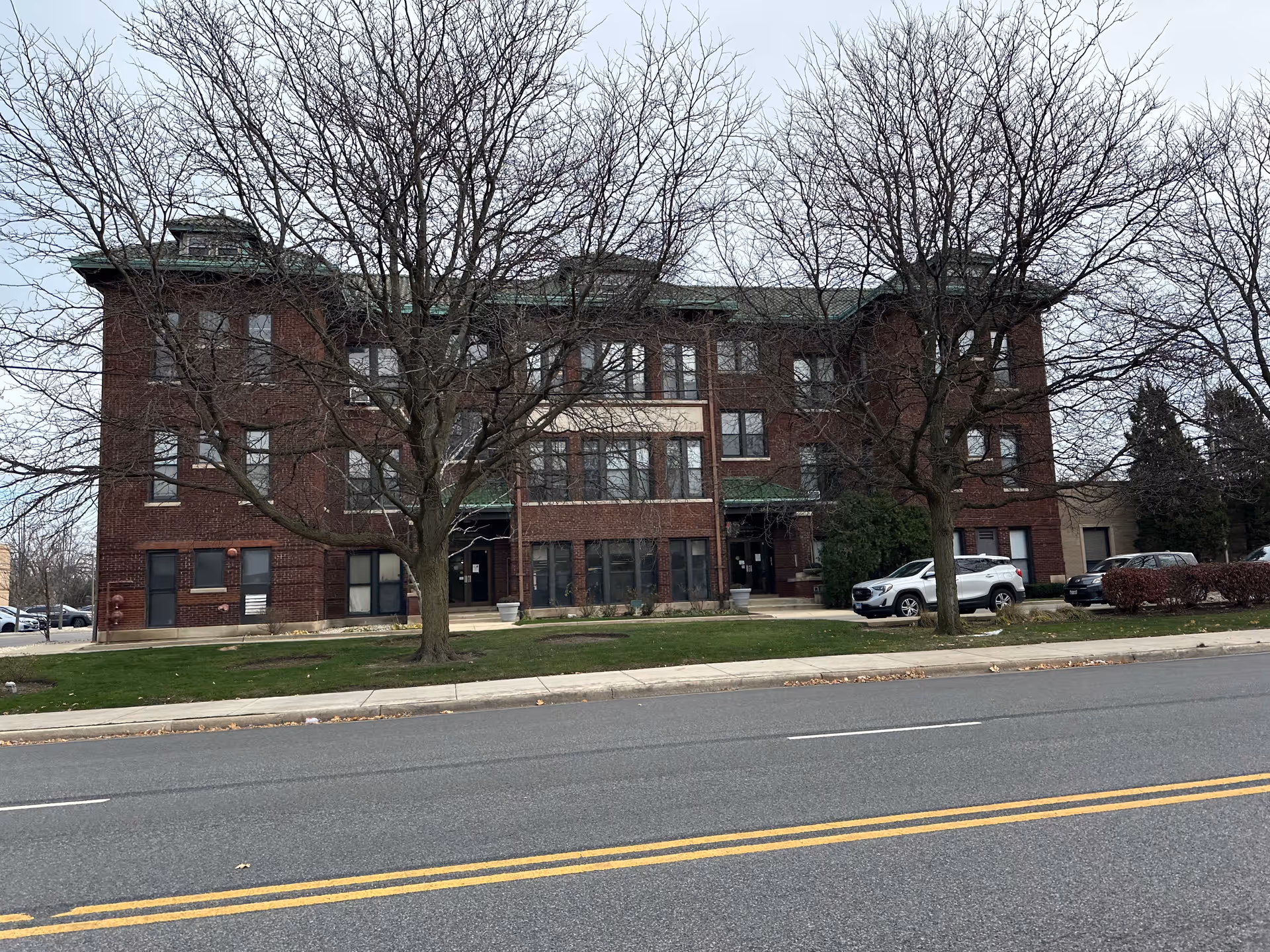 A three-story brick building with multiple windows and two leafless trees in front. There is a green lawn and a sidewalk separating the building from a paved road with yellow dividing lines. Several parked cars are visible on the right side of the building.