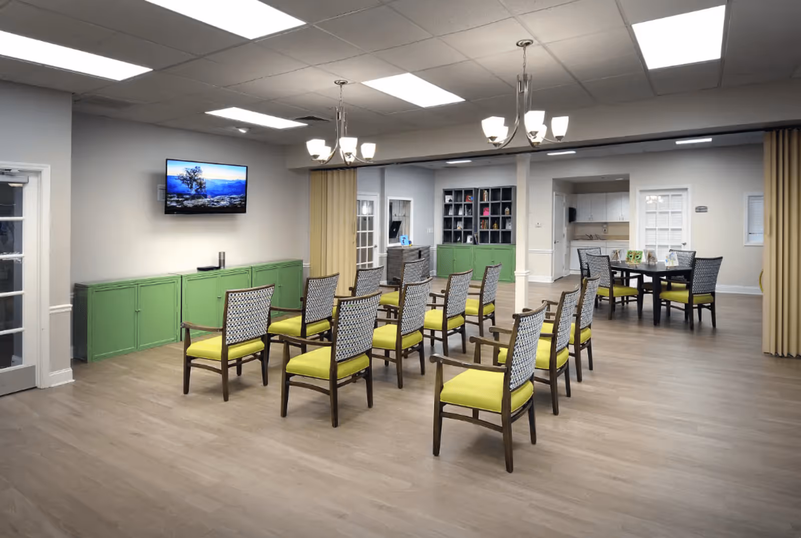 A spacious room with multiple chairs arranged in rows facing a wall-mounted TV. The chairs have wooden frames with patterned backs and bright yellow cushions. The room has light-colored walls, green cabinets along one wall, and two chandeliers hanging from the ceiling. There is also a round table with chairs in the background near a kitchenette area.