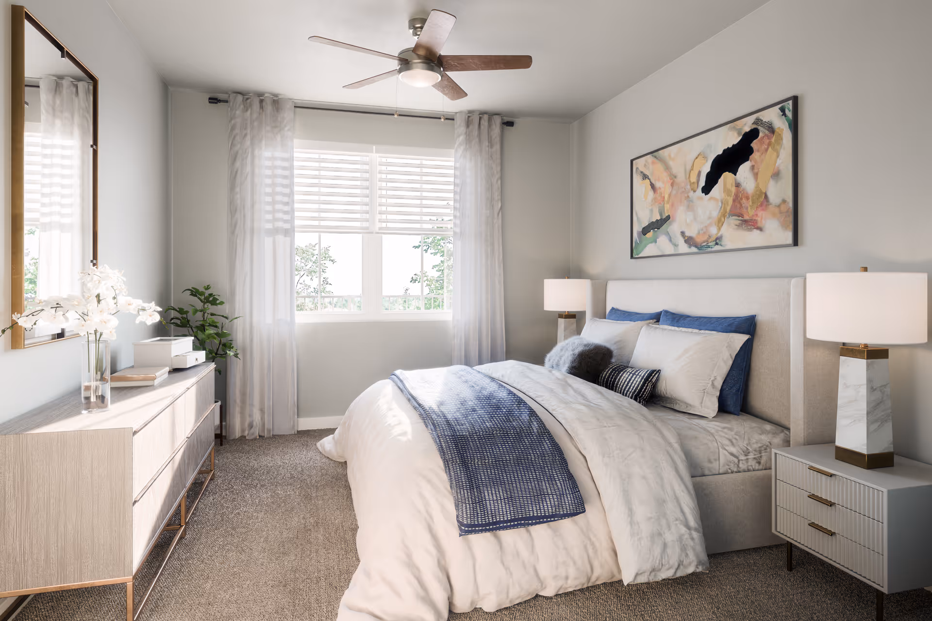 Sunlit, contemporary bedroom featuring a neatly made bed with blue accents, bedside tables and lamps, a dresser under a mirror, a window with sheer curtains, and a ceiling fan.