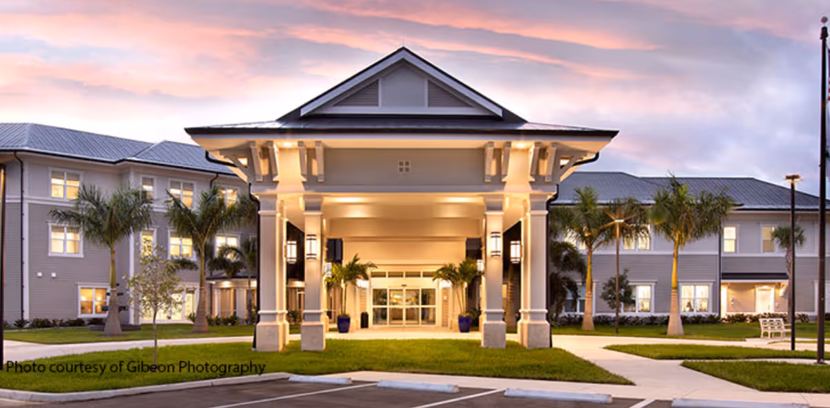 Front entrance of a senior living building with a covered porte-cochère, palm trees, and a colorful evening sky.