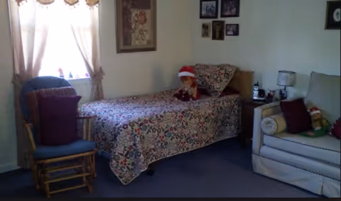 A small assisted-living bedroom with a single bed covered by a floral quilt, a couch, and a rocking chair near a curtained window.