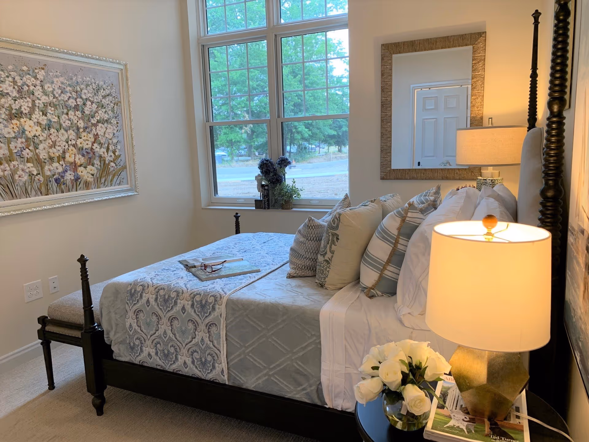 A cozy bedroom with a dark wooden bed frame, a neatly made bed with multiple decorative pillows and a patterned bedspread. A bedside table holds a lamp with a beige shade and a vase of white roses. A large window with a view of trees lets in natural light. A framed floral painting hangs on the wall to the left, and a large mirror is mounted on the wall behind the bed.