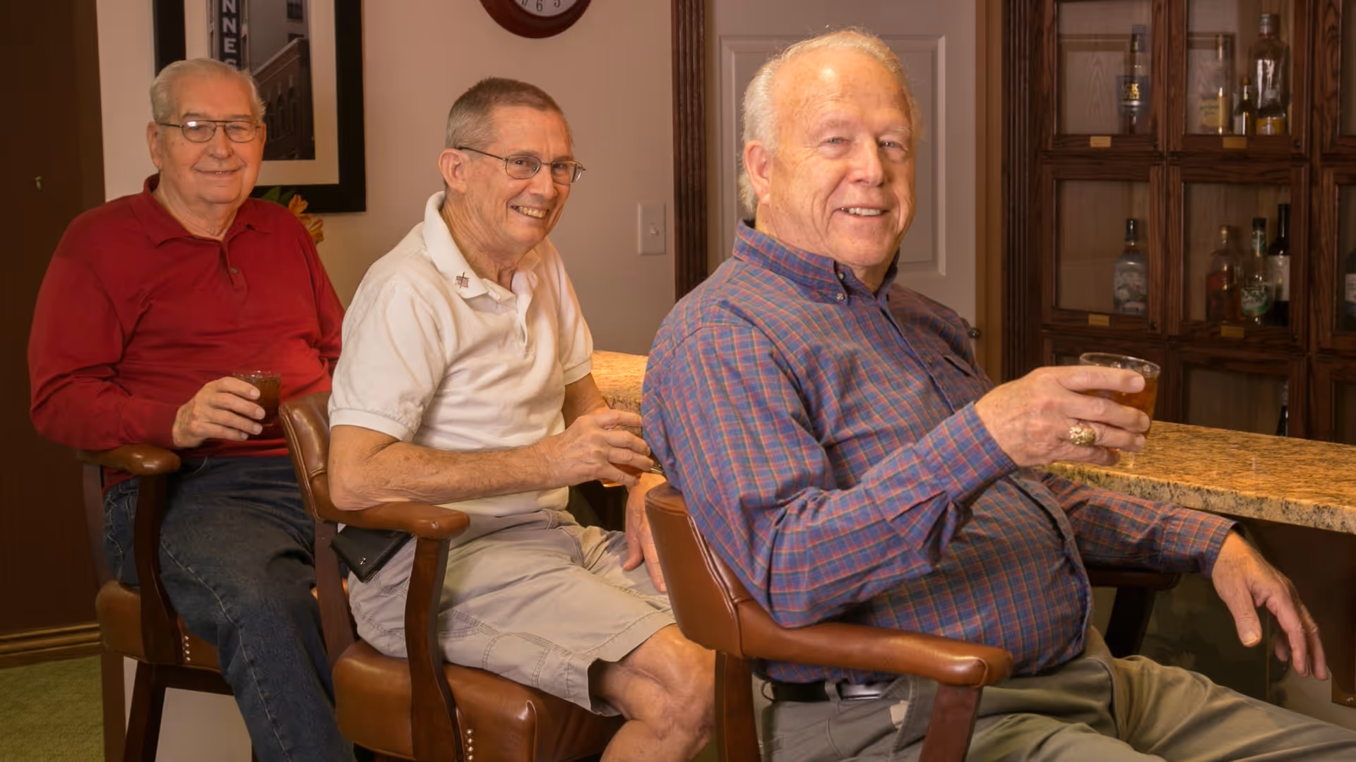 Three elderly men sitting on bar stools at a counter, each holding a drink and smiling. Behind them is a wooden cabinet with various bottles displayed.