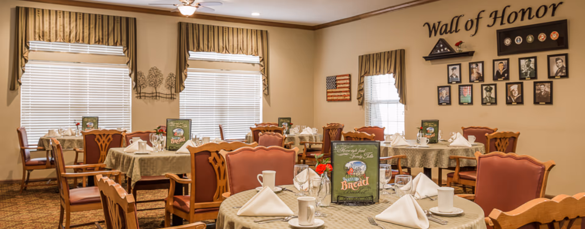 A dining room with multiple round tables covered with beige tablecloths, each set with white napkins, cups, glasses, and menus. The room has wooden chairs with red cushions, two large windows with striped valances, and a wall decorated with a 'Wall of Honor' display featuring framed photos and military insignia. There is also a small American flag decoration on the wall.
