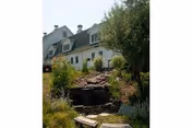 White multi-story building with dormer windows behind a landscaped garden and small stone waterfall.