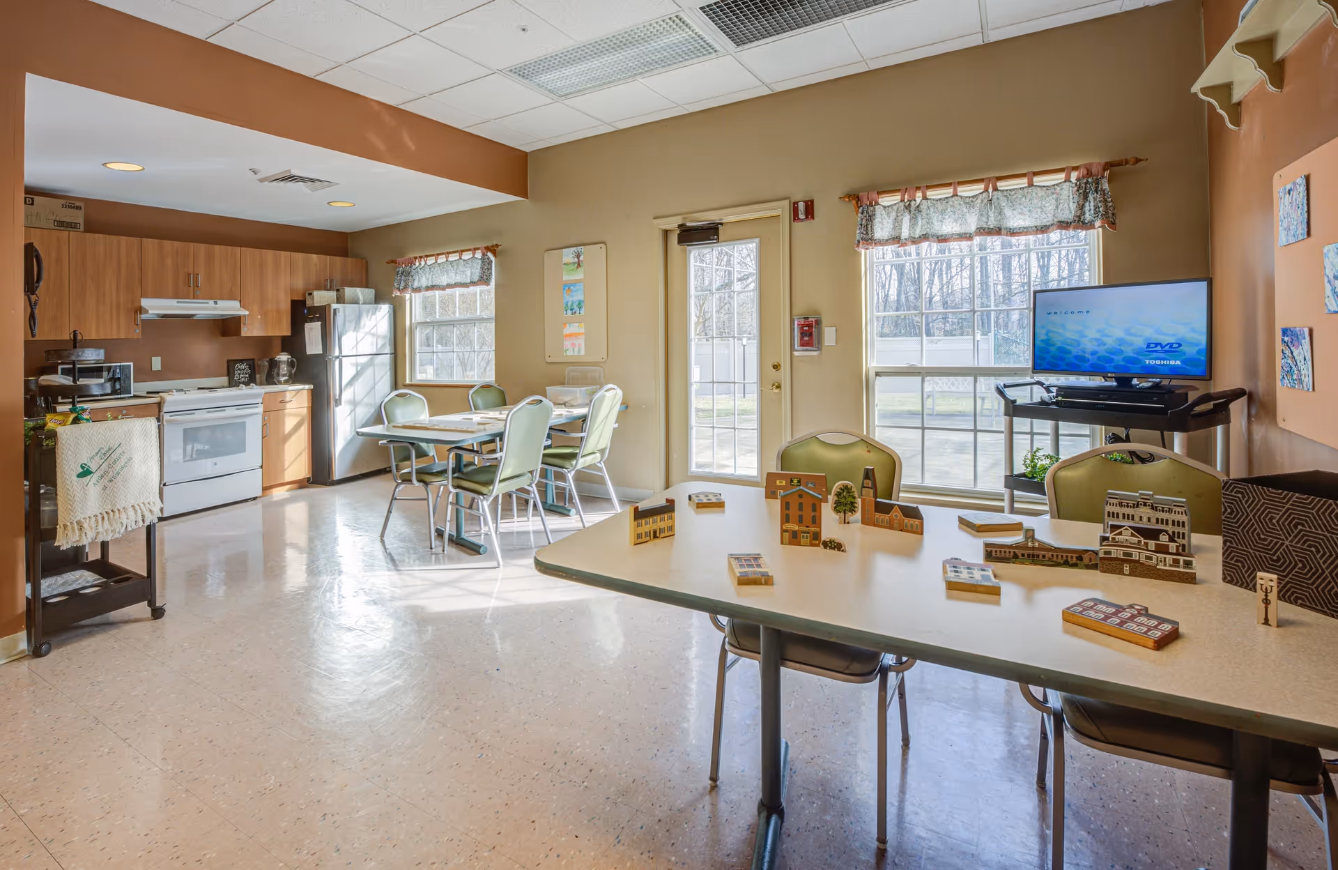 A bright, clean room with a kitchen area featuring wooden cabinets, a white stove, microwave, and stainless steel refrigerator. There are two tables with green chairs around them, one table has small wooden model buildings and blocks on it. A TV on a stand is positioned near two large windows with floral valances, and a door leads outside. The room has beige walls and a tiled floor.