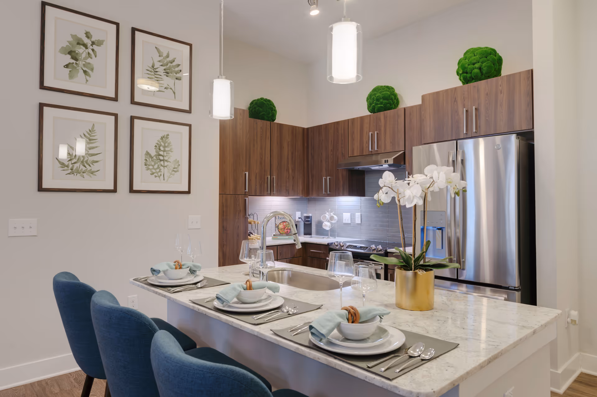 Modern kitchen with a marble countertop island set for four with blue chairs, white plates, bowls, silverware, and light blue napkins. The kitchen features dark wood cabinets, stainless steel refrigerator, stove, and a sink with a curved faucet. There are four framed botanical prints on the wall and three green decorative moss balls on top of the cabinets. Two pendant lights hang from the ceiling.