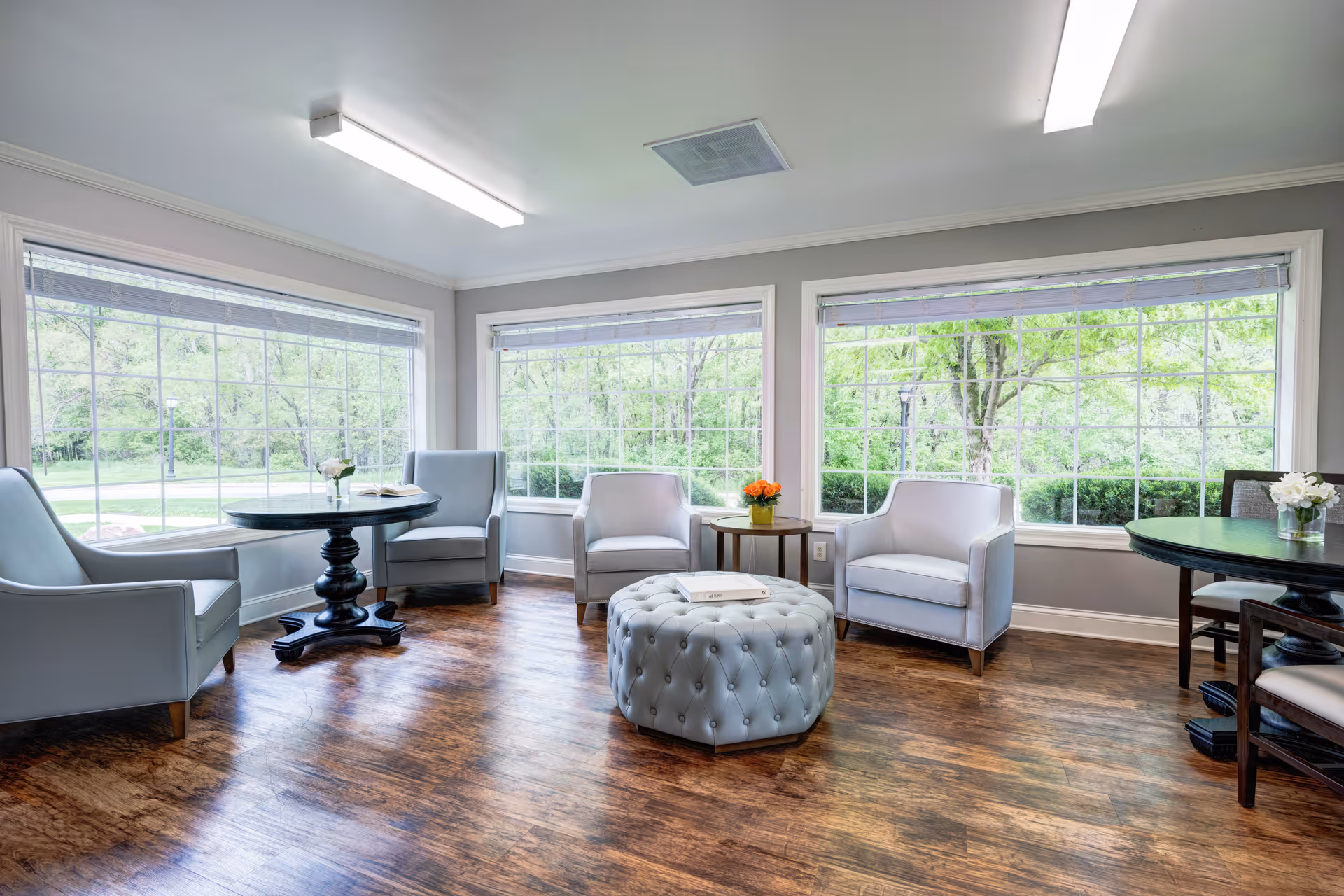 A bright and spacious common area with large windows showing green trees outside. The room features light gray armchairs arranged around a tufted round ottoman in the center. There are two round tables with chairs, each decorated with small flower arrangements. The floor is wooden, and the walls are painted light gray.