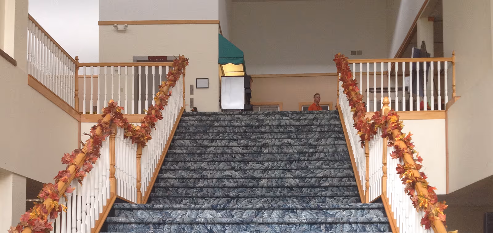 Wide staircase with blue patterned carpet and white wooden railings decorated with autumn leaf garlands on both sides, leading up to a landing with a person standing in the background inside a building.