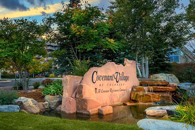 Outdoor landscaped area with a large stone sign that reads 'Covenant Village of Colorado, A Covenant Retirement Community' surrounded by greenery, rocks, and a small water feature with cascading water.