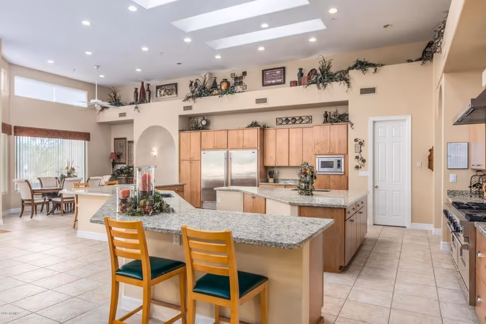 Spacious kitchen area with granite countertops and wooden cabinets. Two wooden chairs with green cushions are placed at the kitchen island. Stainless steel double-door refrigerator and built-in microwave are visible. The kitchen opens into a dining area with a round table and chairs near large windows with blinds. The room is decorated with plants and framed pictures on a high shelf near the ceiling.