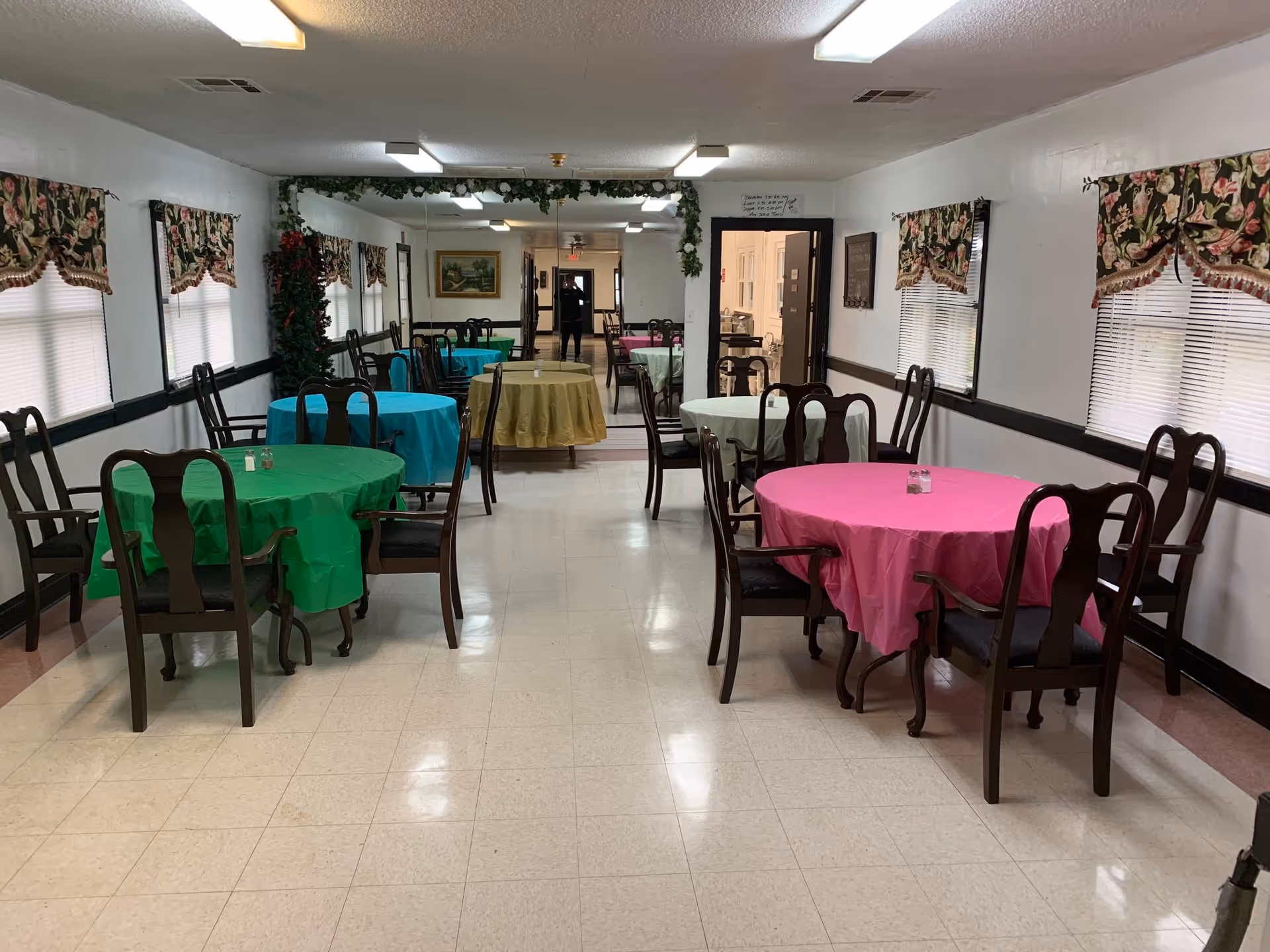 Dining/activity room with several round tables covered in colorful tablecloths and wooden chairs.