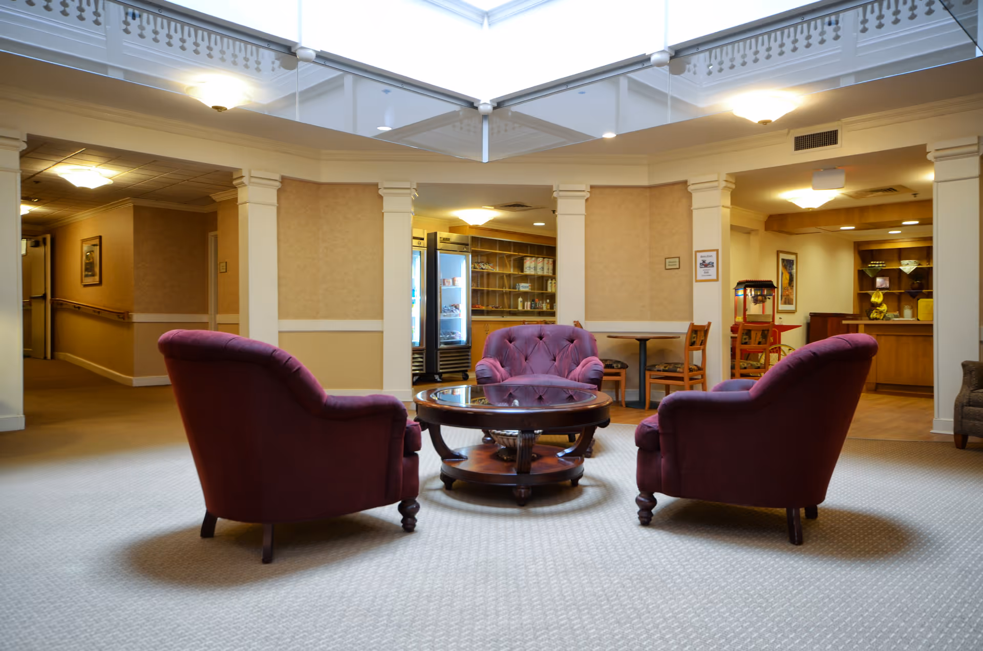 A cozy seating area in a senior living community featuring three plush purple armchairs arranged around a round wooden coffee table. The space is well-lit with ceiling lights and has beige walls with white trim. In the background, there is a small dining area with tables and chairs, a refrigerator, and a popcorn machine.