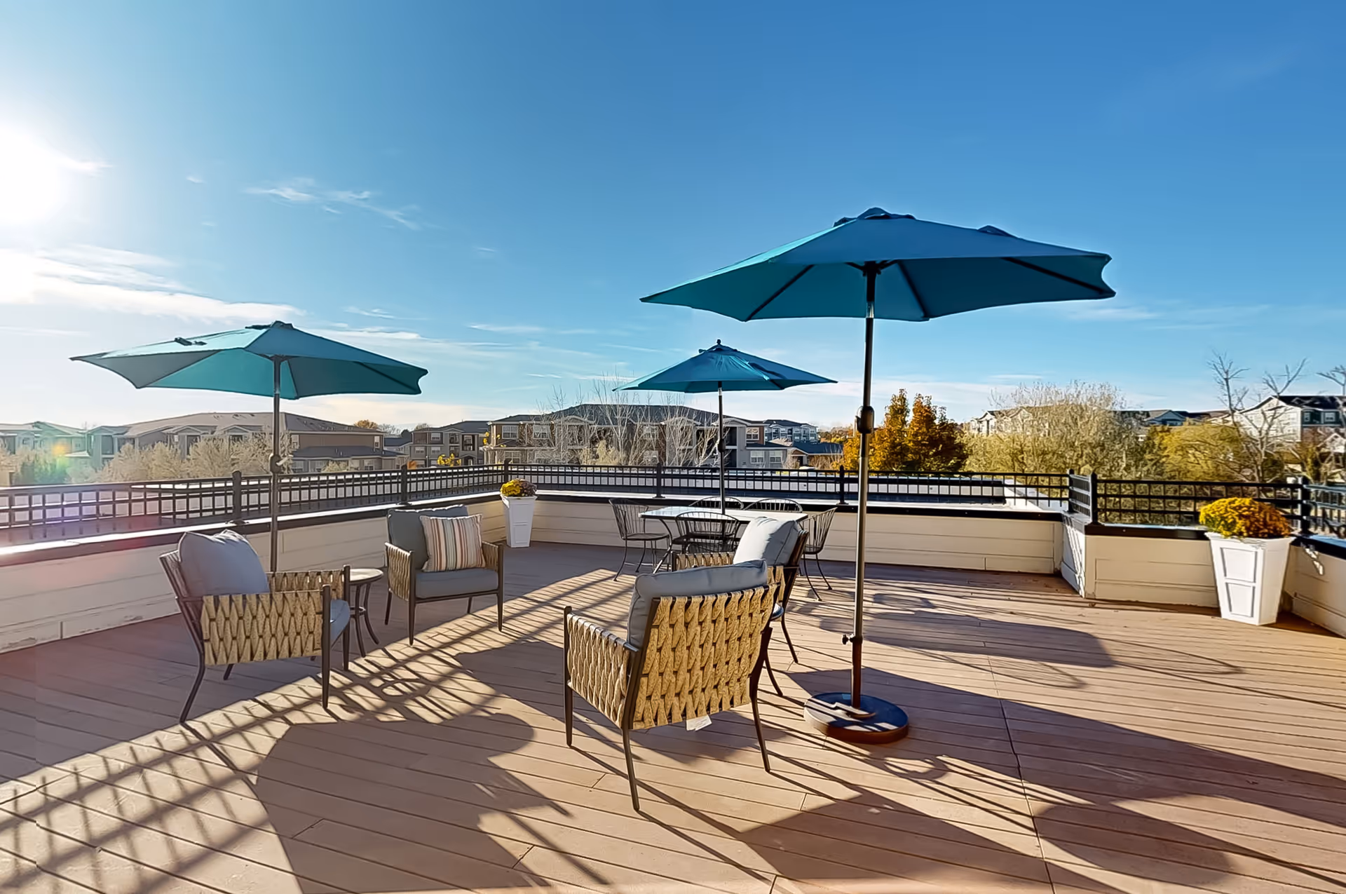 Outdoor patio area with wooden flooring, featuring several seating arrangements including cushioned chairs and tables with blue umbrellas. The patio is surrounded by a railing and overlooks residential buildings and trees under a clear blue sky with the sun shining brightly.